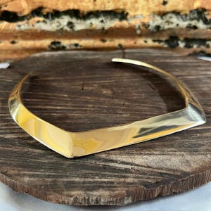 Può includere: Una collana a girocollo color oro con un design a forma di V. La collana ha una finitura lucida ed è esposta su una superficie di legno scuro. La collana è un pezzo forte.