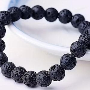 Puede incluir: Una pulsera circular compuesta por numerosas cuentas esféricas de lava negra. Cada cuenta presenta una superficie porosa y texturizada característica de la roca volcánica. Las cuentas están ensartadas en un bucle continuo y uniforme.