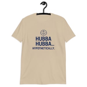 Puede incluir: Camiseta color beige con el texto azul marino "Hubba Hubba... Hypothetically" y un gráfico de cerebro. La camiseta está colgada en una percha negra.