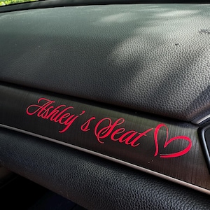 May include: A red decal with the words "Ashley's Seat" in a cursive font, accompanied by a heart symbol. The decal is placed on a dark, textured surface, likely a car's dashboard.