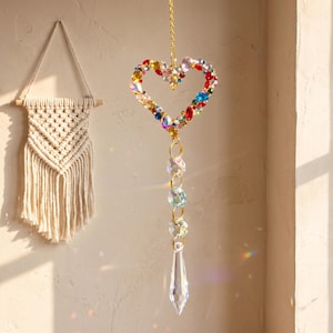 Op de afbeelding: Een hangende zonnevanger met een hartvormig frame ingelegd met veelkleurige gefacetteerde kristallen, opgehangen aan een goudkleurige ketting. Onder het hart verbinden drie heldere bolvormige kristallen zich met een langwerpig druppelprisma.