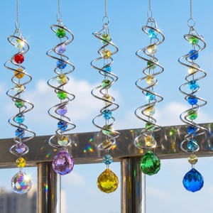 May include: Five hanging spiral wind spinners made of silver-toned metal wire, each featuring a vertical strand of multi-colored faceted glass beads and a large, round, colored crystal pendant at the bottom, reflecting light against a blue sky.