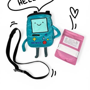 May include: A teal climbing chalk bag in the shape of a cartoon character, with a black strap and a pink chalk bag. The chalk bag has a smiling face and the word "HELLO!" in a speech bubble.