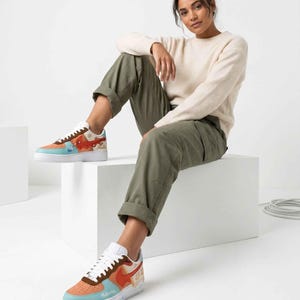May include: Customized sneakers with a colorful design. The shoes feature a white sole, a light blue toe, and a brown and orange upper with artwork. The word "Moana" is visible on the side. The shoes are worn by a person.