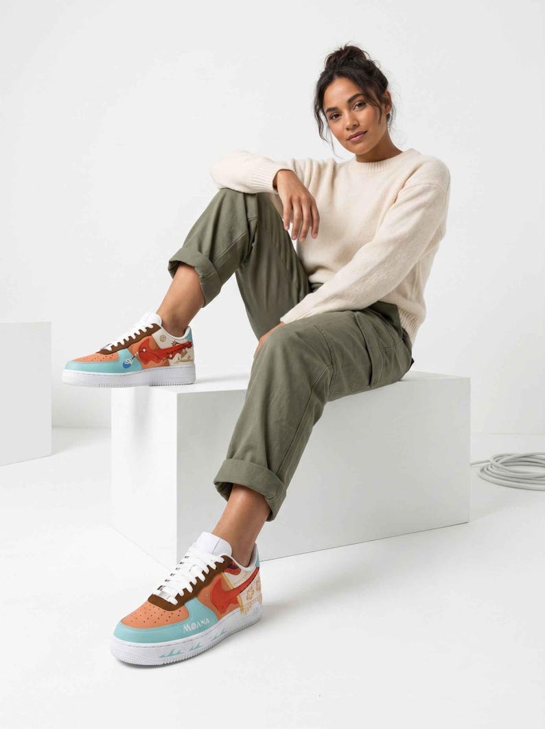 May include: Customized sneakers with a colorful design. The shoes feature a white sole, a light blue toe, and a brown and orange upper with artwork. The word "Moana" is visible on the side. The shoes are worn by a person.