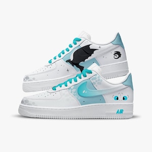 May include: White sneakers with light blue accents and laces. One shoe features a black dragon graphic, while the other has a light blue swoosh and a cartoon dragon face. The shoes have the word "AIR" on the side.