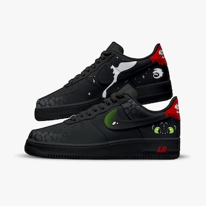 May include: Black trainers with custom dragon-themed artwork. One shoe features a white dragon design, while the other showcases a green-eyed dragon face. The trainers have a black swoosh and the word "AIR" in red.
