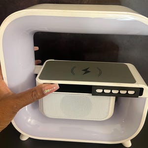 May include: A white and gray bedside lamp with a wireless charging pad and speaker. The lamp has a curved design with a light strip and a control panel. The charging pad has a lightning bolt symbol. The speaker has a white mesh cover.