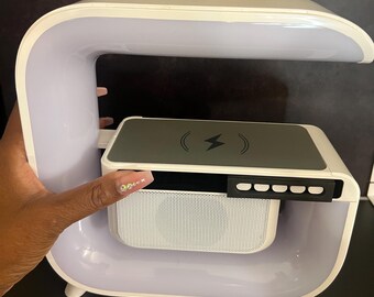 Puede incluir: Una lámpara de noche blanca y gris con una plataforma de carga inalámbrica y un altavoz. La lámpara tiene un diseño curvo con una tira de luz y un panel de control. La plataforma de carga tiene un símbolo de rayo. El altavoz tiene una cubierta de malla blanca.