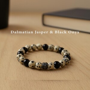 Könnte beinhalten: Ein Armband mit Perlen aus Dalmatiner-Jaspis und schwarzem Onyx. Das Armband kombiniert gesprenkelte beige und schwarze Perlen. Der Text "Dalmatian Jasper & Black Onyx" wird über dem Armband angezeigt.