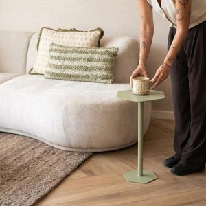 Könnte beinhalten: Ein hellgrüner sechseckiger Beistelltisch mit einem einzelnen zentralen Bein. Eine strukturierte beige Tasse steht auf dem Tisch. Der Tisch steht neben einem beigen Sofa mit grünen und beigen Kissen. Der Boden ist aus Holz.