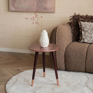 Könnte beinhalten: Kleiner, runder Beistelltisch mit kupferfarbener Tischplatte und dunkelbraunen Beinen. Die Beine haben kupferfarbene Spitzen. Eine weiße Vase mit rosa Blumen steht auf dem Tisch. Der Tisch steht auf einem weißen Teppich.