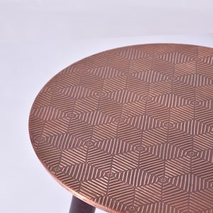 May include: A round copper-colored side table with a geometric pattern on the tabletop. The tabletop has a repeating design of hexagons and lines. The table has a dark brown leg.