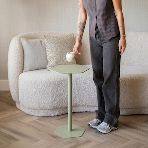Puede incluir: Una mesa auxiliar hexagonal verde salvia con un único soporte central y una base circular. La mesa mide aproximadamente 71 cm de altura. La mesa está junto a un sofá blanco curvado con cojines estampados. El suelo es de madera marrón claro.