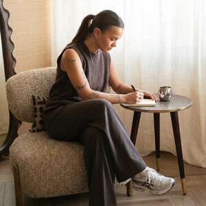 May include: A person seated in a textured armchair, writing in a notebook at a small round table. The table has a patterned top and dark wooden legs. A silver cup sits on the table. The person is wearing a brown sleeveless top and trousers, with white trainers.