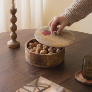 Peut inclure: Un bol rond en bois avec un couvercle assorti orné d'une illustration de grenade rouge. Le bol est rempli de noix brunes et possède un petit bouton argenté sur le couvercle. Le grain du bois est visible sur toute la surface du récipient.