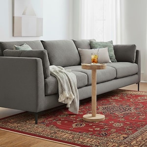 Könnte beinhalten: Ein graues Stoffsofa mit mehreren Kissen und einer hellen Decke. Ein kleiner Beistelltisch aus Holz trägt ein Glas Eistee. Das Sofa steht auf einem roten gemusterten Teppich.