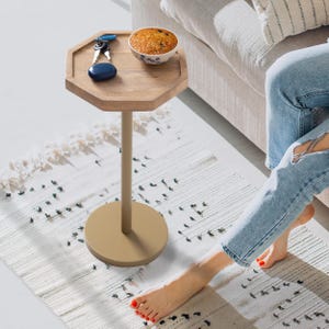 Könnte beinhalten: Kleiner, achteckiger Beistelltisch aus Holz mit beigefarbenem Metallsockel. Auf der Tischplatte liegen Schlüssel, ein blaues ovales Objekt und eine kleine Schüssel mit Backwaren. Der Tisch steht neben einem Sofa und einem gemusterten Teppich.