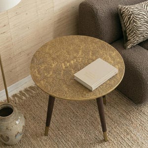 Könnte beinhalten: Ein runder Beistelltisch mit einer goldfarbenen Tischplatte, die ein geprägtes florales Damastmuster aufweist, und drei dunkelbraunen, konisch zulaufenden Holzbeinen mit goldfarbenen Metallfüßen. Ein geschlossenes, helles Buch liegt auf der Tischplatte.
