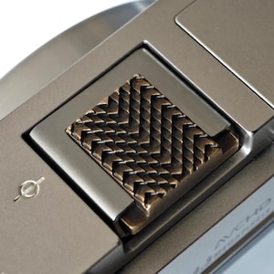 May include: Close-up of a silver digital camera with a textured bronze-colored grip. The camera body has a metallic finish and features a small dial and the text "X 5000".