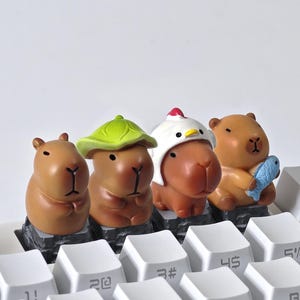 May include: Four brown capybara figurines, each with unique accessories, sit atop a white keyboard. One wears a green leaf hat, another a chicken hat, and the last holds a blue fish. The keyboard keys are white.