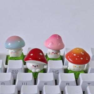 May include: Four mushroom-shaped keycaps in various colours, including blue, pink, red, and orange, sit on a white keyboard. Each keycap features a smiling face. The keyboard keys display numbers and letters.