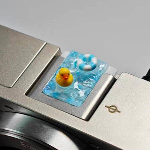 May include: A silver camera with a decorative resin piece featuring a yellow rubber duck and a blue and white life preserver floating in a pool of water. The resin piece is on the top of the camera.