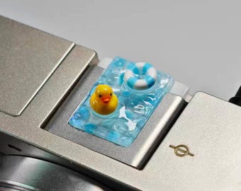 May include: A silver camera with a decorative resin piece featuring a yellow rubber duck and a blue and white life preserver floating in a pool of water. The resin piece is on the top of the camera.