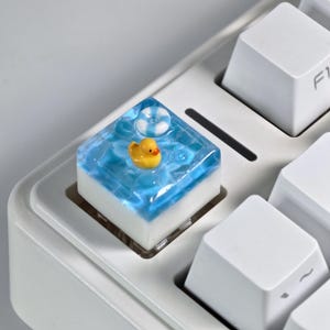 Könnte beinhalten: Eine individuelle Tastaturkappe mit einer gelben Gummiente, die in einer blauen Wasserszene schwimmt, mit einer kleinen weiß-blauen Schwimmboje. Die quadratische Kappe befindet sich auf einer weißen Tastatur.
