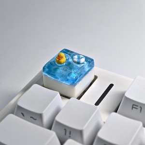 May include: A custom keyboard keycap featuring a miniature yellow rubber ducky and a white and blue life preserver floating in a blue water scene. The keycap is set on a white keyboard, with other white keys visible.