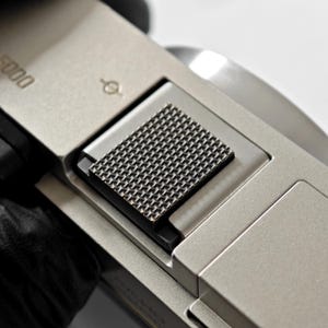 May include: Close-up of a silver-toned camera body with a textured, square-shaped grip. The grip is made of a dark, patterned material. The camera has the number "5000" printed on it.