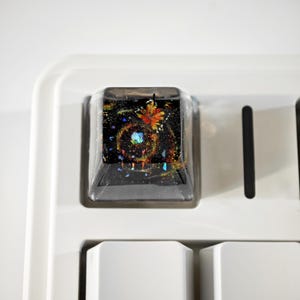 May include: Close-up of a custom keyboard keycap with a black base and a clear, resin-filled top. The resin contains a colorful, galaxy-like design with orange, blue, and yellow hues. The keycap is set on a white keyboard.