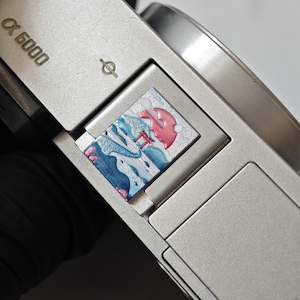 Puede incluir: Una tapa para zapata de cámara de color plateado con una incrustación de esmalte cuadrada. El diseño muestra un paisaje estilizado con colinas azules, un torii rojo y un sol rosa sobre un fondo blanco. El cuerpo de la cámara tiene la marca Alpha 6000.