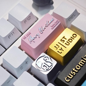 Może przedstawiać: Zbliżenie klawiatury mechanicznej z niestandardowymi nakładkami na klawisze. Różowa nakładka na klawisz głosi "Merry Christmas" i "Love you Vanemax". Złota nakładka na klawisz głosi "JJ ST LY UDIO". Inne klawisze są białe z etykietami funkcji.