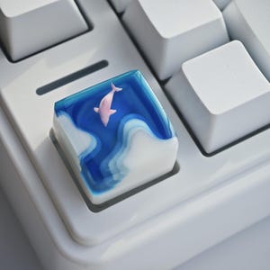 May include: A custom keycap featuring a pink dolphin swimming in a blue and white ocean design. The keycap is square and sits on a white keyboard. The dolphin is the focal point, with the ocean waves adding depth.