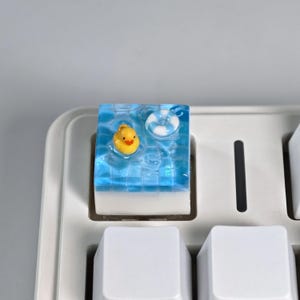 May include: A custom keyboard keycap featuring a miniature pool scene. The keycap has a clear, blue resin top with a yellow rubber duck and a white and blue life preserver floating inside. The keycap is mounted on a white keyboard.
