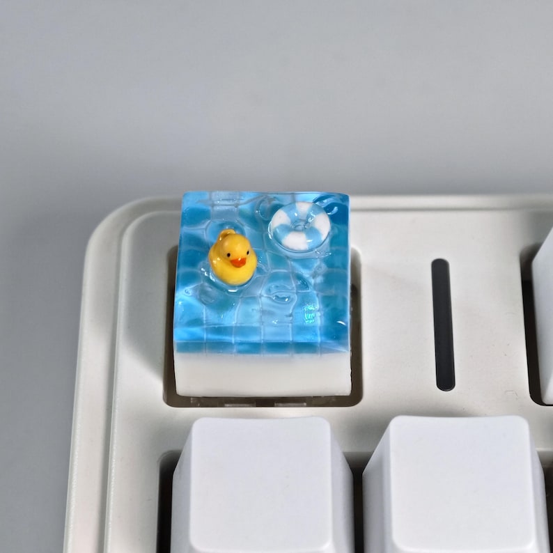 May include: A custom keyboard keycap featuring a miniature pool scene. The keycap has a clear, blue resin top with a yellow rubber duck and a white and blue life preserver floating inside. The keycap is mounted on a white keyboard.