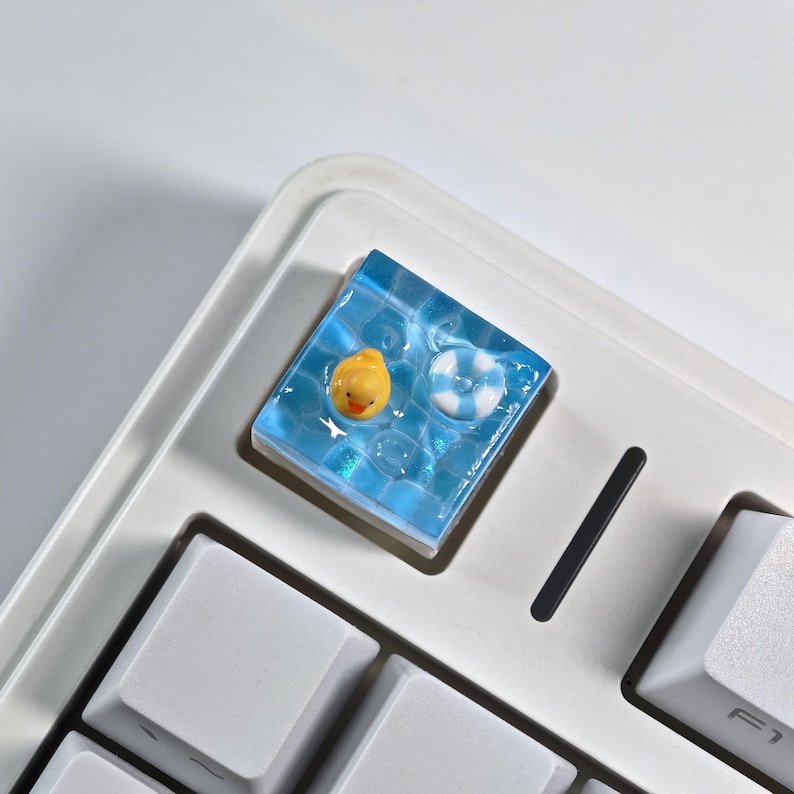 May include: A custom keycap featuring a miniature pool scene. The keycap has a blue, water-like background with a yellow rubber duck and a white and blue life preserver. The keycap is square and sits on a white keyboard.