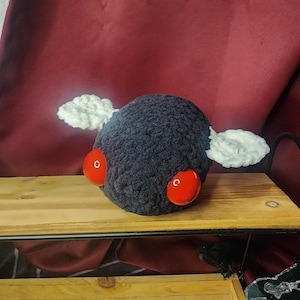 May include: A handmade, dark grey crocheted creature with large red eyes and two white cloud-like wings. The plush toy is on a wooden shelf against a burgundy background. The toy has a round body.