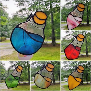 May include: A collection of stained glass potion bottle suncatchers, each featuring a black leaded frame, a yellow cork stopper, and a unique colored glass body in blue, pink, red, green, white, or yellow, suspended by a black string for display.