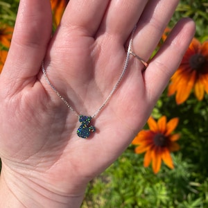 May include: A silver necklace featuring a small, dark blue and green bear-shaped pendant. The pendant has an iridescent sheen. The necklace is held in a hand, set against a background of green grass and orange flowers.