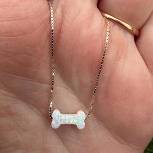 May include: A delicate silver chain necklace featuring a bone-shaped pendant. The pendant is white with iridescent flecks, giving it a shimmering appearance. The necklace is a simple, elegant piece of jewelry.
