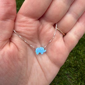 May include: A silver necklace featuring a small, light blue opal elephant pendant. The elephant pendant has a subtle, iridescent glow. The necklace is displayed against a background of a hand and green grass.
