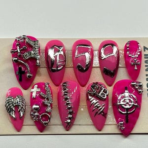 May include: A set of ten hot pink press-on nails with pointed tips, featuring silver-toned metallic charms including crosses, wings, chains, and numbers. The nails are displayed on a rectangular card labeled RRRADICALNAILZ on both sides.