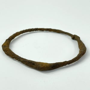 May include: A weathered, circular metal bracelet with a twisted design. The bracelet has a rusty brown color and appears to be antique or vintage. The metal is textured and shows signs of age and wear, suggesting it is an old piece of jewelry.