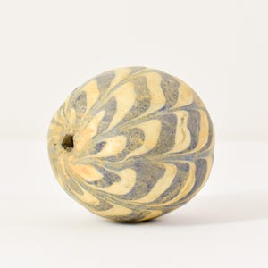 Op de afbeelding: Een ronde, gemarmerde kraal met een gat. De kraal heeft een wervelend patroon van crème- en blauwgrijze kleuren. De kraal is een decoratief item, mogelijk voor sieraden of ambachten.