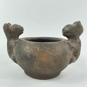 May include: A handcrafted, brown ceramic bowl with two animal figures on either side. The bowl has a wide opening and a rounded base. The animal figures appear to be stylized cats or similar creatures, adding a unique decorative element.