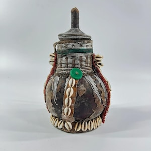 May include: A rounded gourd vessel with a pointed lid, decorated with white cowrie shells, red and white beadwork, silver-toned metallic threads, and a single green button. The surface features a dark, weathered texture with visible patches.