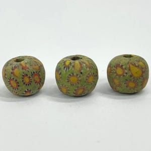 May include: Three round, olive-green beads with a floral pattern in yellow, red, and black. The beads have a hole running through the center. The beads are likely made of glass and are suitable for jewelry making or other crafts.