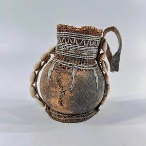 May include: A small, round, brown gourd vessel featuring a textured neck wrapped in silver-colored wire detailing. The exterior is adorned with a vertical row of white cowrie shells along the side and a sturdy, dark brown leather handle.